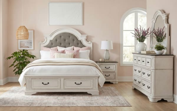 Bedroom set featuring a white wooden bed frame with tufted gray upholstered headboard, matching nightstand and dresser with mirror, soft pink walls, and decorative plants.